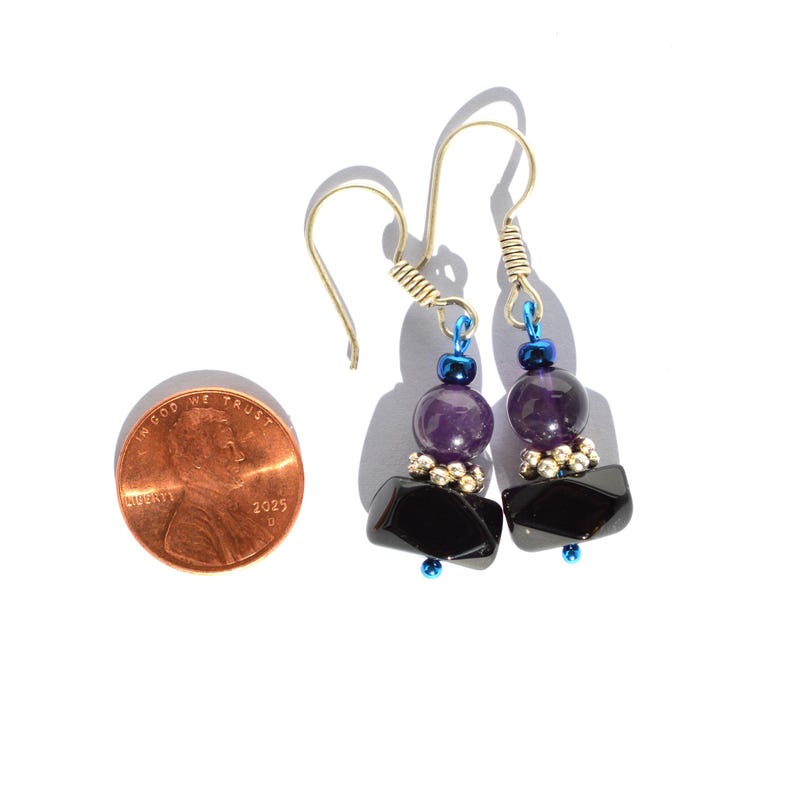 May include: A pair of dangle earrings with a combination of black, purple, and silver beads. The earrings have a silver hook and a blue accent bead. A U.S. penny is next to the earrings for size reference.