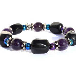 May include: A beaded bracelet featuring alternating black and purple gemstones. The bracelet includes small silver-colored beads and iridescent blue beads. The gemstones are polished and have a smooth texture.