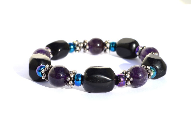 May include: A beaded bracelet featuring alternating black and purple gemstones. The bracelet includes small silver-colored beads and iridescent blue beads. The gemstones are polished and have a smooth texture.