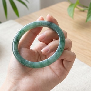 May include: A smooth, circular jade bangle bracelet featuring a translucent green color with darker green mottled inclusions throughout the stone. The bangle has a polished, rounded surface and a consistent thickness around the entire circumference.