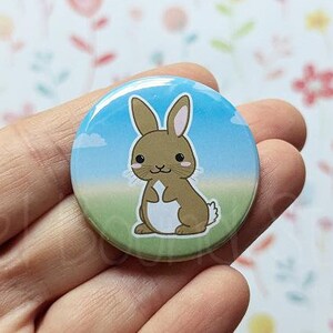 May include: A round button with a cartoon brown bunny rabbit with white accents standing on a green field with a blue sky and white clouds in the background.