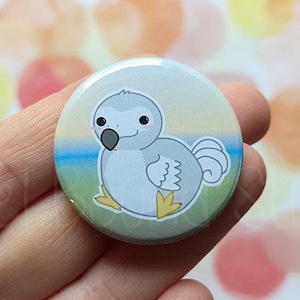 Könnte beinhalten: Ein runder Knopf mit einer Cartoon-Illustration eines grauen Dodos mit gelben Füßen vor einem blauen und grünen Hintergrund. Der Knopf hat einen Durchmesser von 2,5 cm.
