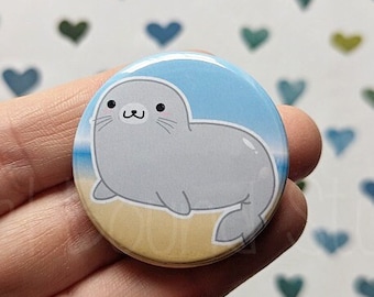 May include: A round button with a cartoon seal on a beach. The seal is gray with a light blue background. The button is 1 inch in diameter.