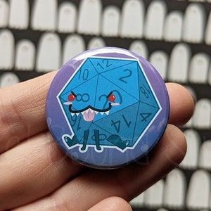 May include: A blue and purple button with a cartoon illustration of a blue 20-sided die with red eyes, a toothy grin, and a tongue sticking out. The die is standing on two legs and has a black shadow underneath. The button has a white background with a pattern of white ghosts.
