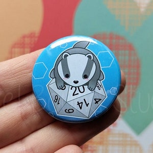 May include: A blue and white button featuring a cartoon badger sitting on top of a twenty-sided die. The die has numbers 4, 6, 8, 10, 12, 14, 16, 18, 20 printed on its faces.
