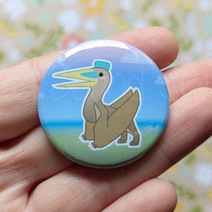 Peut inclure: Un bouton rond avec une illustration de dessin animé d'un ptérosaure brun portant un chapeau bleu et jaune. Le ptérosaure se tient sur une plage avec un ciel bleu et des nuages en arrière-plan.