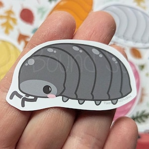 May include: A die-cut sticker featuring a cartoon illustration of a gray pill bug with a rounded segmented body, small black eyes, and tiny pink cheeks. The sticker has a white border and is held against a background of colorful autumn leaves.