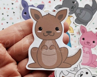 May include: A set of six colorful kangaroo stickers with cute cartoon faces. The stickers are on a white background with a pink, green, and gray triangle pattern.