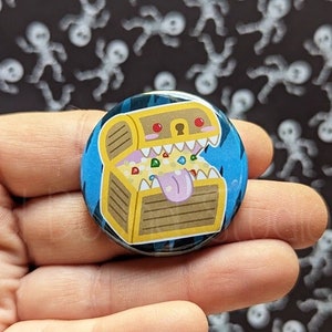 May include: A blue and yellow button with a cartoon image of a treasure chest with a tongue sticking out. The chest is filled with colorful gems and gold coins.