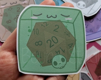 May include: A hand holding several stickers featuring a D20 die inside a clear cube with a skull and kawaii face. The stickers are in various colors, including green, blue, and pink, with the text "Pixel Bound Studios".