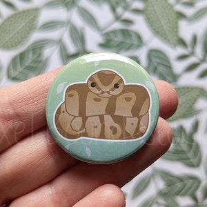 May include: A round button featuring a green background with a brown snake curled up on it. The snake has a light brown head and a darker brown body.