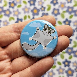 May include: A blue button with a cartoon sugar glider flying against a blue sky with white clouds. The sugar glider is gray with black eyes and a black nose.