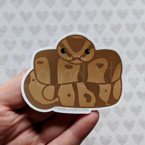 May include: A brown and tan snake curled up in a ball, with a cute cartoon face. The sticker has the text "PixelBoundStudios" at the bottom.