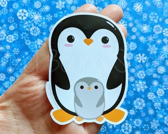 以下が含まれることがあります： 黒と白のペンギンと、その中にいる小さな灰色のペンギンのステッカー。ペンギンはオレンジ色のくちばしと足を持っています。ステッカーは、白い雪片が散りばめられた青い背景に貼られています。