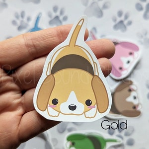 May include: A gold vinyl sticker of a cartoon beagle dog with brown ears and a brown collar. The dog is looking down with its tongue sticking out. The sticker is on a white background with a paw print pattern.
