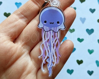 May include: A clear acrylic keychain featuring a cartoon jellyfish with a purple body, white eyes, and pink and white tentacles. The keychain is held in a hand against a background of blue and green hearts.
