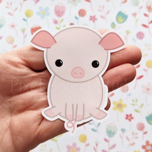 Puede incluir: Una pegatina de cerdo rosa claro con ojos negros, un hocico rosa y orejas rosas. El cerdo está sentado con la cola enroscada. El fondo es un estampado floral. La pegatina se sostiene en una mano. El texto de la imagen dice "Pixel Bound Studios."