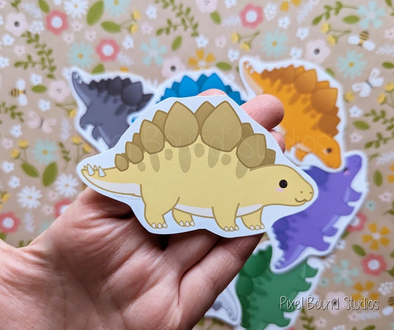 Puede incluir: Un conjunto de pegatinas de dinosaurios de colores que incluyen un estegosaurio amarillo y marr&oacute;n, un estegosaurio azul, un estegosaurio morado, un estegosaurio verde, un estegosaurio gris y un estegosaurio naranja. Las pegatinas est&aacute;n sobre un fondo floral.