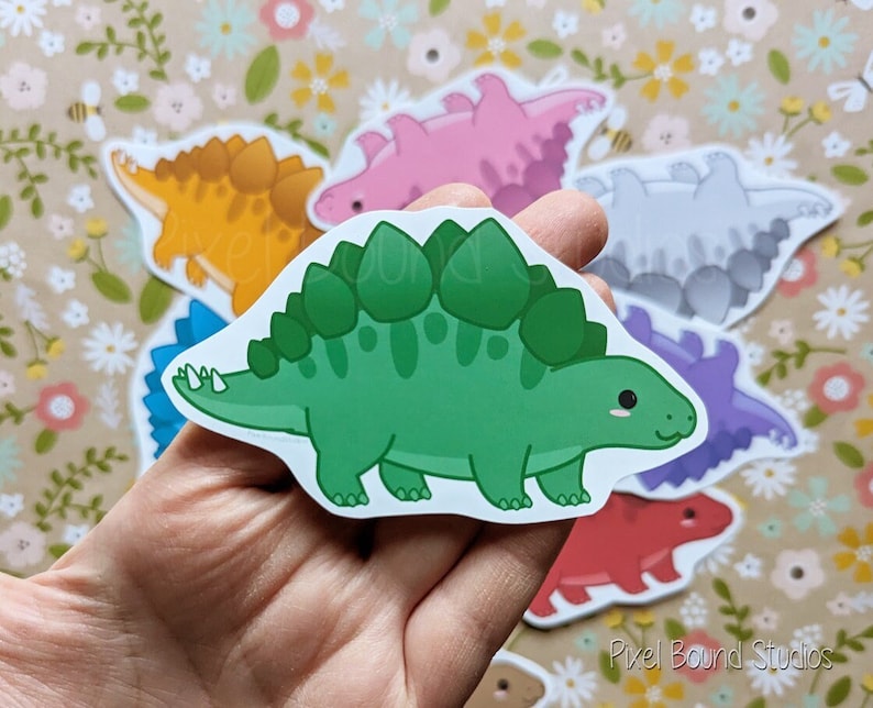 Puede incluir: Un conjunto de nueve pegatinas de dinosaurios de colores que representan diferentes especies, incluyendo un estegosaurio verde, un braquiosaurio rosa, un anquilosaurio amarillo, un paquicefalosaurio gris, un parasaurolophus morado y un triceratops rojo. Las pegatinas est&aacute;n sobre un fondo floral.