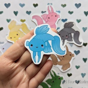 Pode incluir: Um conjunto de adesivos de peixes coloridos com um peixe betta azul, um axolotl rosa, um peixe dourado amarelo, um peixe-anjo preto e branco e um guppy castanho e branco. Os adesivos est&atilde;o em um fundo branco com cora&ccedil;&otilde;es azuis e verdes.