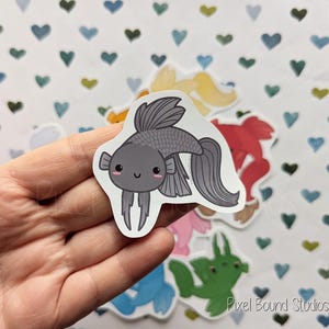 Pode incluir: Um adesivo de peixe de desenho animado cinza e preto com um rosto bonito. O adesivo est&aacute; em uma m&atilde;o contra um fundo de cora&ccedil;&otilde;es brancos.