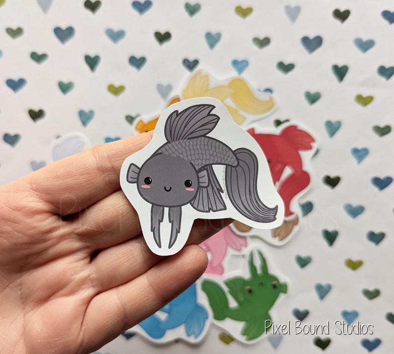 Pode incluir: Um adesivo de peixe de desenho animado cinza e preto com um rosto bonito. O adesivo est&aacute; em uma m&atilde;o contra um fundo de cora&ccedil;&otilde;es brancos.
