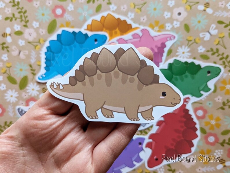 Puede incluir: Un conjunto de seis pegatinas de dinosaurios de colores que representan diferentes especies, incluyendo un estegosaurio marr&oacute;n, un anquilosaurio azul, un paquicefalosaurio rosa, un estegosaurio verde, un anquilosaurio rojo y un paquicefalosaurio morado. Las pegatinas est&aacute;n sobre un fondo con estampado floral.