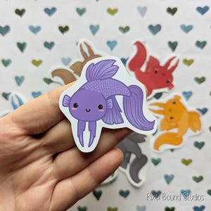 Pode incluir: Um adesivo de peixe de desenho animado roxo e branco com um rosto sorridente. O adesivo est&aacute; em uma m&atilde;o com um fundo de cora&ccedil;&otilde;es.