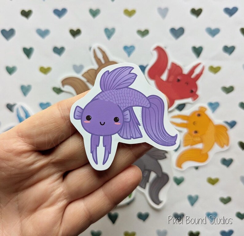 Pode incluir: Um adesivo de peixe de desenho animado roxo e branco com um rosto sorridente. O adesivo est&aacute; em uma m&atilde;o com um fundo de cora&ccedil;&otilde;es.