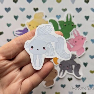 Pode incluir: Um adesivo de peixe branco e azul com um rosto de desenho animado fofo. O adesivo est&aacute; em uma m&atilde;o contra um fundo de cora&ccedil;&otilde;es e outros adesivos.