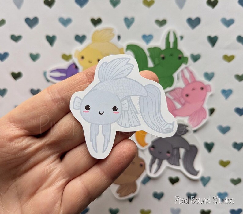 Pode incluir: Um adesivo de peixe branco e azul com um rosto de desenho animado fofo. O adesivo est&aacute; em uma m&atilde;o contra um fundo de cora&ccedil;&otilde;es e outros adesivos.