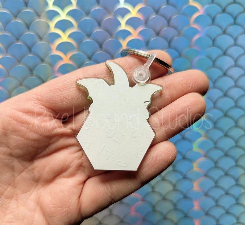 May include: White acrylic keychain shaped like a Dungeons and Dragons die with a 4-sided design. The keychain has a silver metal ring attached.