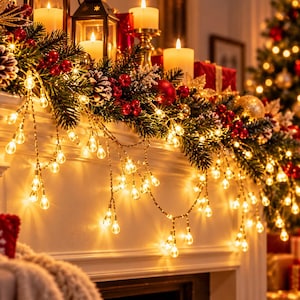 Puede incluir: Una cadena de luces LED de color blanco cálido con bombillas en forma de lágrima sobre una repisa de chimenea decorada con ramas de pino verdes, bayas rojas, piñas, adornos dorados y velas encendidas en farolillos de cristal.