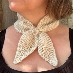 May include: A cream-colored, hand-knitted neckerchief featuring a textured garter stitch pattern. The scarf is designed to be tied in a knot at the front, creating two pointed ends that drape downwards. The yarn has a soft, natural off-white tone.