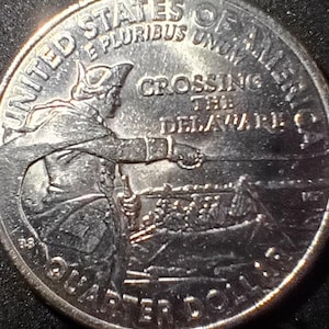 May include: A silver quarter coin featuring an image of George Washington leading a boat across the Delaware River. The coin has the text "UNITED STATES OF AMERICA", "E PLURIBUS UNUM", "CROSSING THE DELAWARE", and "QUARTER DOLLAR".