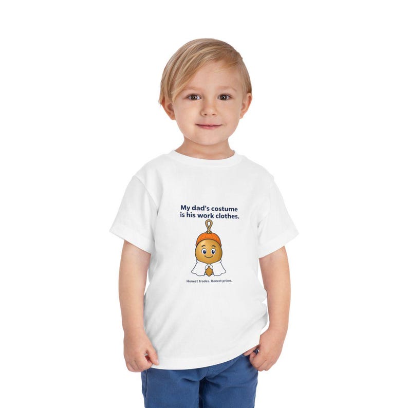 May include: A white short-sleeved t-shirt featuring a graphic of a cartoon acorn wearing a ghost costume. The text reads: My dad's costume is his work clothes. Honest trades. Honest prices. The shirt is made of a plain white fabric material.