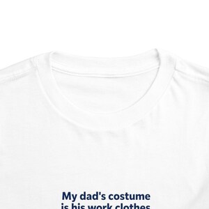 May include: A white crew neck t-shirt featuring dark blue text printed on the front. The text reads, My dad's costume is his work clothes. The garment has a standard ribbed collar and short sleeves, showcasing a clean and simple design.