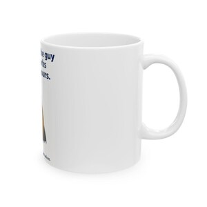 May include: A white ceramic mug featuring a graphic of a person in a dark cape and the text: I am the guy who works long hours. The design includes dark blue lettering and a stylised figure with a brown and black cape on the side of the mug.