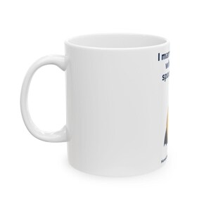 May include: A white ceramic mug featuring a dark blue text graphic that reads I married a witch and she spotted me. Below the text is a stylised orange pumpkin illustration with a black witch hat on top. The mug has a curved handle on the left side.