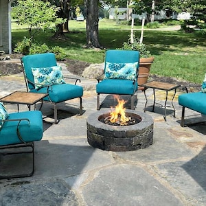 May include: A patio set consisting of three teal armchairs with dark metal frames and matching teal cushions. Each chair features a decorative throw pillow with a blue and white floral pattern. A circular stone fire pit sits in the center.