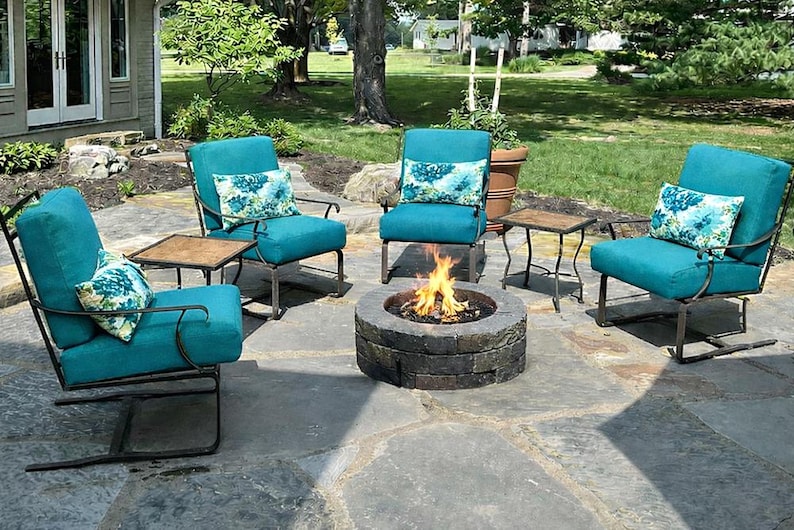 May include: A patio set consisting of three teal armchairs with dark metal frames and matching teal cushions. Each chair features a decorative throw pillow with a blue and white floral pattern. A circular stone fire pit sits in the center.