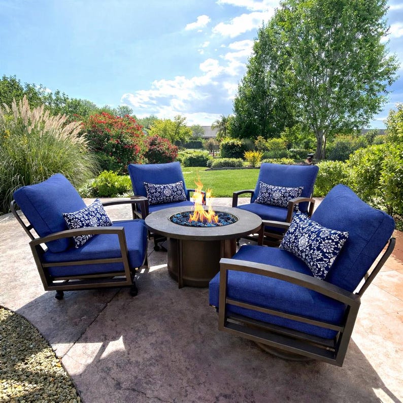 May include: A set of four dark blue outdoor armchairs with metal frames and matching blue cushions, each featuring a small rectangular white and blue patterned lumbar pillow, arranged around a circular bronze-colored gas fire pit table.