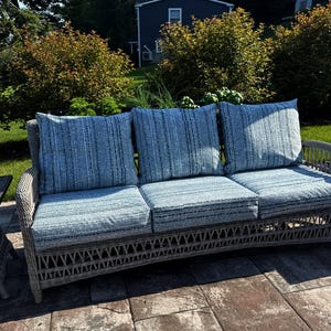 May include: A three-seat outdoor sofa featuring a light gray woven wicker frame and three thick seat cushions paired with three matching back cushions, all upholstered in a blue and white textured fabric with a subtle vertical striped pattern.