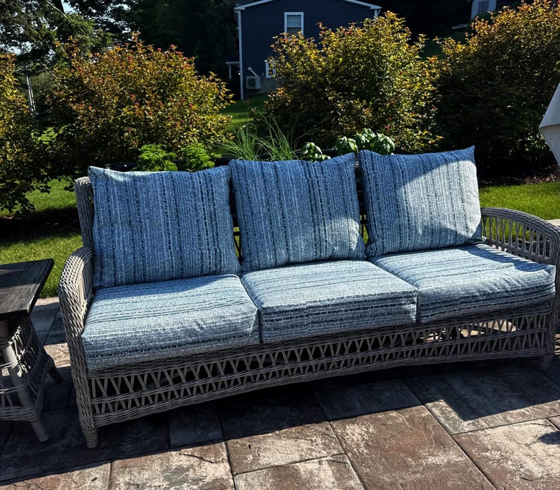 May include: A three-seat outdoor sofa featuring a light gray woven wicker frame and three thick seat cushions paired with three matching back cushions, all upholstered in a blue and white textured fabric with a subtle vertical striped pattern.