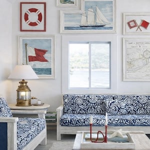 May include: A living room set featuring a sofa and armchair with white and blue patterned cushions, a brass nautical lamp, and a gallery wall displaying framed artwork of lighthouses, sailboats, signal flags, and a nautical chart on white walls.