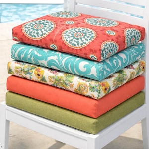 May include: A stack of five outdoor seat cushions in various colors and patterns. The top cushion is coral with teal and white circular designs. Other cushions are teal, floral, coral, and green. The cushions are stacked on a white chair.