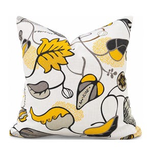 May include: A square throw pillow featuring a white background with a repeating botanical pattern of stylized leaves and vines in shades of mustard yellow, charcoal gray, and black, accented with small dotted textures throughout.