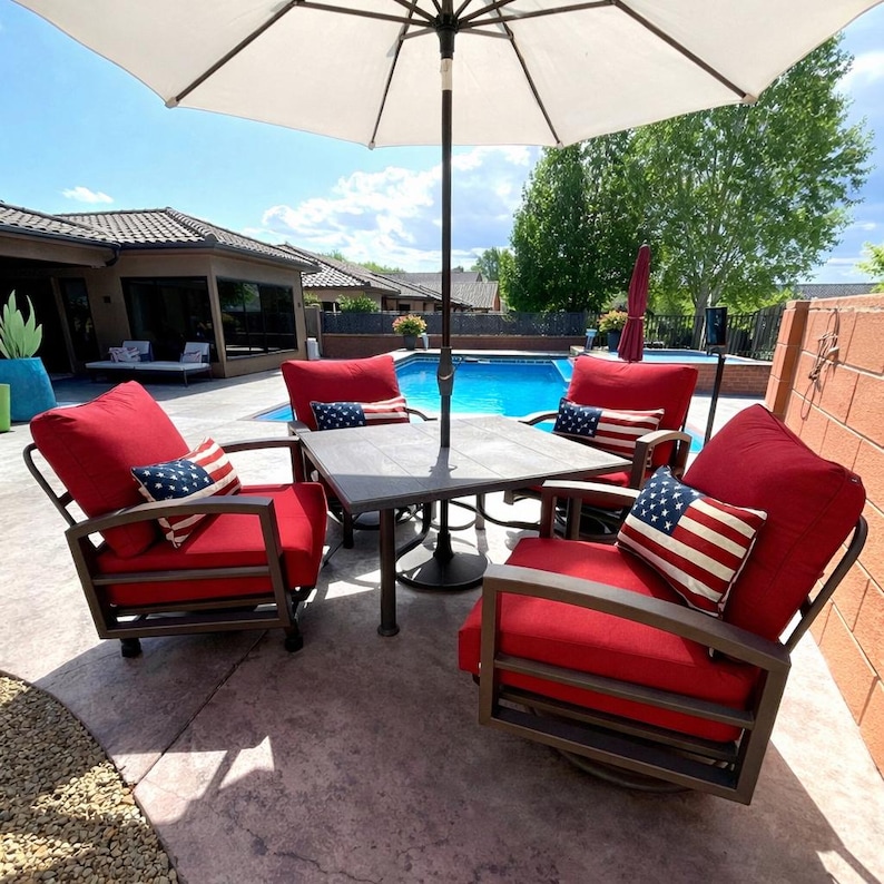 May include: A patio dining set featuring a square dark metal table with a central umbrella hole, surrounded by four matching metal chairs with thick red cushions and rectangular throw pillows displaying the American flag pattern.