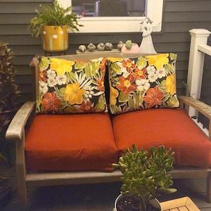 May include: A wooden loveseat featuring two solid burnt orange seat cushions and two square throw pillows. The pillows display a vibrant tropical floral pattern with yellow, orange, and white flowers set against a dark green and black foliage background.