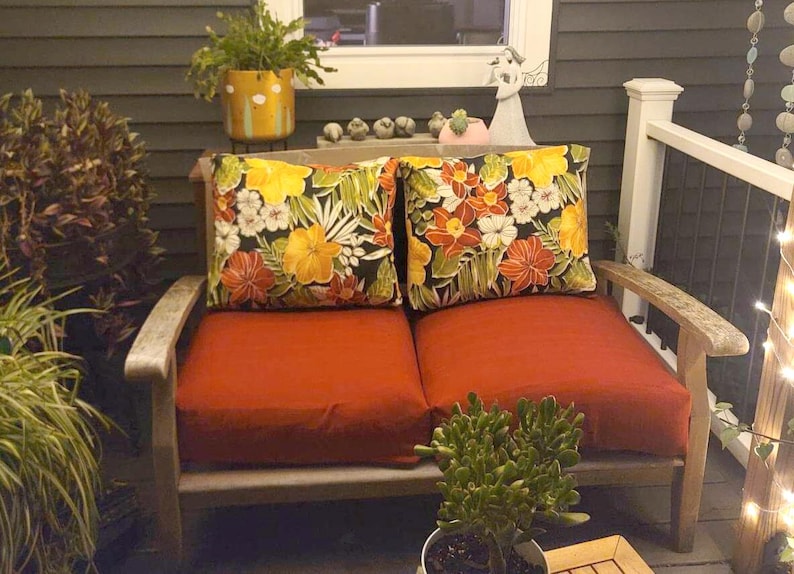 May include: A wooden loveseat featuring two solid burnt orange seat cushions and two square throw pillows. The pillows display a vibrant tropical floral pattern with yellow, orange, and white flowers set against a dark green and black foliage background.
